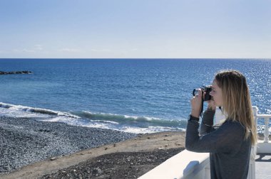 Bir kamera tutarak ve okyanus, çakıl plaj ve sahil bir güneşli sabah Tenerife, Kanarya Adaları, İspanya'da karşı karşıya bir fotoğraf alarak beyaz elbise genç bin yıllık erkek