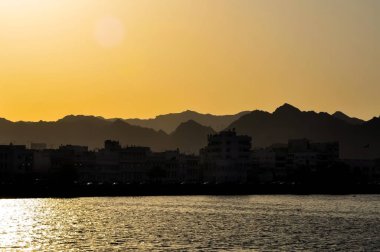 Dağların silueti ve Mutrah limanı civarındaki şehir binaları, Umman.