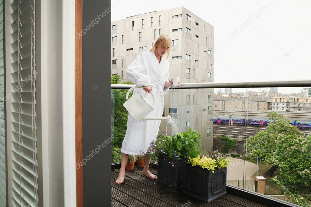 attractive young caucasian woman in bathrobe watering flowers on balcony