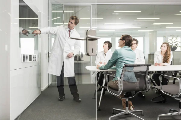 Hombre que lleva abrigo de laboratorio apuntando a la pantalla en la sala de conferencias, cuatro colegas que observan — Foto de Stock