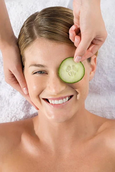 Stock image Smiling young woman lying on massage table with cucumber slice being placed over eye