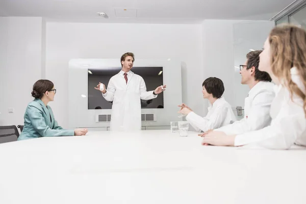 Hombre con abrigo de laboratorio sentado al final de la mesa de la sala de conferencias, cuatro colegas sentados. — Foto de Stock