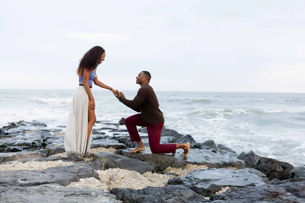 Hombre adulto medio arrodillado sobre rocas junto al mar, proponiéndole matrimonio a una joven — Foto de Stock