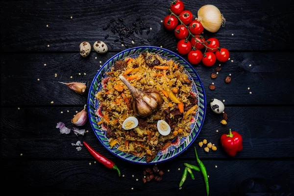 Pilaf und Zutaten auf Teller mit orientalischem Ornament auf dunklem Holzgrund. Zentralasiatische Küche - Plov von oben — Stockfoto