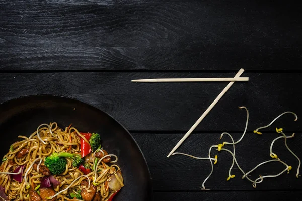 Udon sofríe los fideos con pollo y verduras en una sartén wok sobre fondo de madera negra. Con palillos —  Fotos de Stock