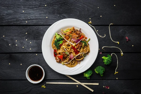Udon stir fry erişte tavuk ve sebze siyah ahşap zemin üzerine beyaz bir plaka ile. Yemek çubukları ve sos ile