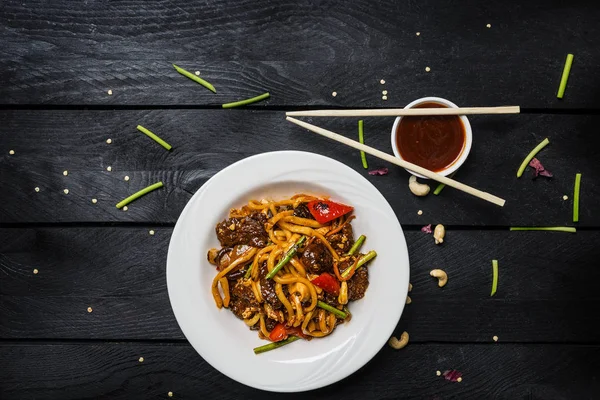 Udon stir fry erişte et ve sebze siyah ahşap zemin üzerine beyaz bir plaka ile. Yemek çubukları ve sos ile — Stok fotoğraf