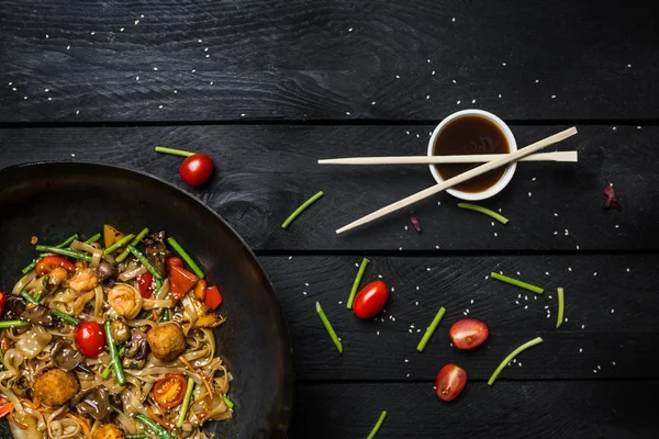 Stir fry nudlích s mořských plodů a zeleniny wok pánvi na černém pozadí dřevěná. Hůlky s omáčkou. — Stock fotografie