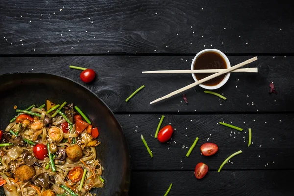 Udon heyecan fry erişte ile deniz ürünleri ve sebze siyah ahşap zemin üzerine wok tavada. Yemek çubukları ve sos ile.