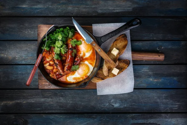 Sıcak bir tavada kızarmış yumurta, sosis, fasulye, yeşil ve tost ile kahvaltı. Bir renkli ahşap arka plan üzerine çatal bıçak takımı ile. Üstten Görünüm