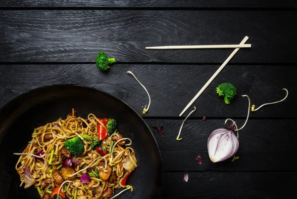 Udon sofríe los fideos con pollo y verduras en una sartén wok sobre fondo de madera negra con palillos. Vista superior —  Fotos de Stock