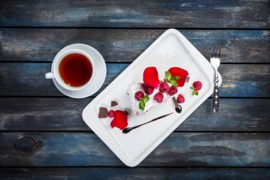 Bir fincan çay lezzetli Pavlova pasta. Gül yaprakları ile beyaz bir plaka üzerinde taze ahududu. Üstten Görünüm. Güzel ahşap arka plan.