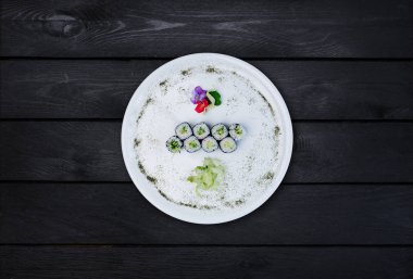 Bir beyaz yuvarlak tabakta klasik suşi roll salatalık ile küçük çiçekler, Japon yemekleri, üstten görünüm ile dekore edilmiştir. Siyah ahşap arka plan