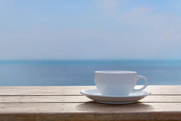 Weiße Kaffeetasse auf Holztischplatte mit verschwommenem Meereshimmel-Hintergrund — Stockfoto