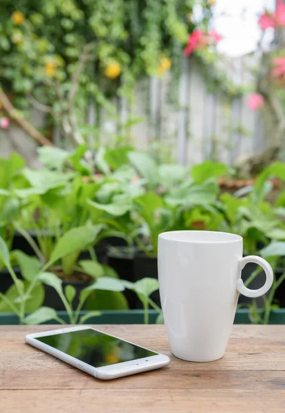野菜畑や花の庭の背景に木製の板の上の携帯電話や白いコーヒーカップ — ストック写真