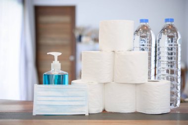 Coronavirus veya COVID-19 nedeniyle ev karantinası el jeli, doku rulosu, içme suyu şişesi ve oturma odasında cerrahi maske ile korunuyor.