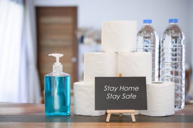 Coronavirus veya COVID-19 nedeniyle ev karantinası el jeli, doku rulosu, içme suyu şişesi ve oturma odasındaki siyah tahta levhası ile korunuyor.