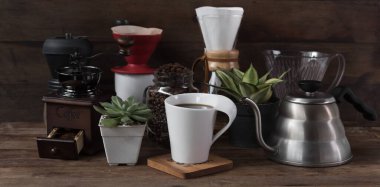 Kahve seti, kavrulmuş fasulye, çaydanlık, öğütücü, beyaz bardak ve çiçek saksısı ahşap masa ve arka planda.