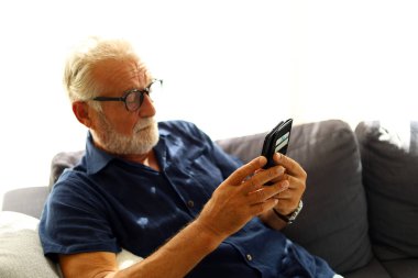 Yaşlı kıdemli adam evde yalnız oturuyor ve cep telefonuyla oynuyor..