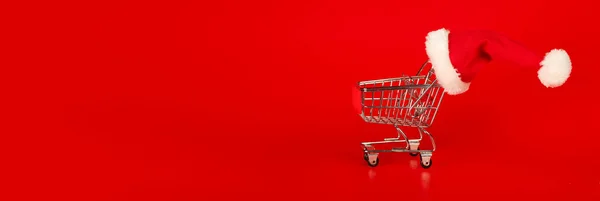 Shopping cart with Christmas Santa hat on red background. Xmas s — Stock Photo, Image