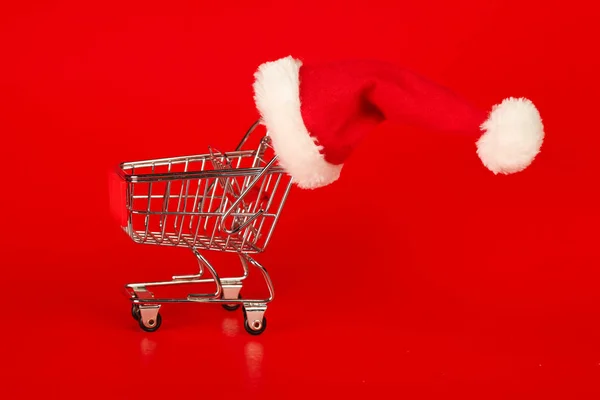 Carrinho de compras com chapéu de Natal Papai Noel no fundo vermelho. Xmas s — Fotografia de Stock