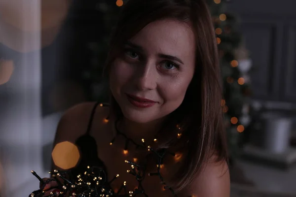 happy young woman with brown hair in a black dress with sequins at the New Year tree with gifts