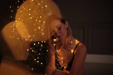 Kısa sarı saçlı güzel bir kız Noel ağacı süslemeli bir Noel ağacı için çelenge sarılmış oturuyor.