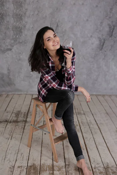 美丽纤细的姑娘 乌黑的短发 身穿勃艮第格子衬衫 喝着一杯红葡萄酒 酒体呈淡灰色 — 图库照片