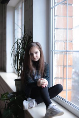 young smiling girl of eight years with long hair in a black turtleneck and jeans sundress walks on the windowsill near the window in a good mood different emotions
