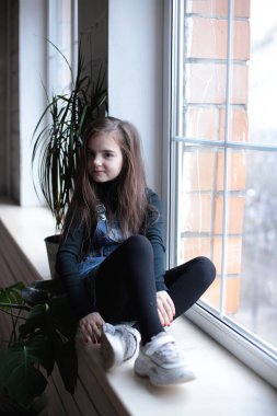 young smiling girl of eight years with long hair in a black turtleneck and jeans sundress walks on the windowsill near the window in a good mood different emotions