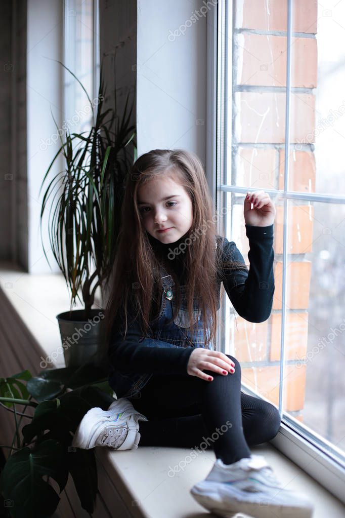 young smiling girl of eight years with long hair in a black turtleneck and jeans sundress walks on the windowsill near the window in a good mood different emotions