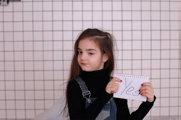 young kind playful girl of eight years with long hair in a black turtleneck and denim sundress with the sign \