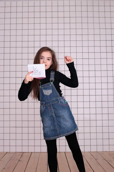 young kind playful girl of eight years with long hair in a black turtleneck and denim sundress with the sign \