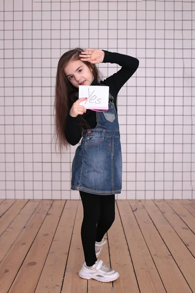 young kind playful girl of eight years with long hair in a black turtleneck and denim sundress with the sign \