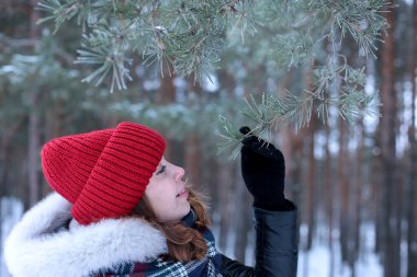 Beautiful girl with green eyes and ginger hair in a red hat on a walk in a winter snowy forest