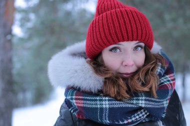Beautiful girl with green eyes and ginger hair in a red hat on a walk in a winter snowy forest