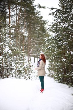 Güzel, çekici, gülümseyen, yeşil gözlü, kızıl saçlı genç bir kadın kışın karlı bir ormanda keyifli bir şekilde yürüyor.