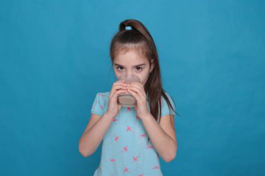 beautiful eight-year-old girl drinks warm milk from a transparent glass on a blue background in a blue t-shirt