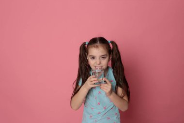 A beautiful girl of eight years drinks water from a transparent glass on a pink background in a blue T-shirt