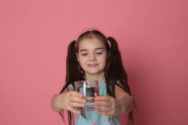 A beautiful girl of eight years drinks water from a transparent glass on a pink background in a blue T-shirt