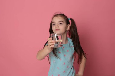 A beautiful girl of eight years drinks water from a transparent glass on a pink background in a blue T-shirt