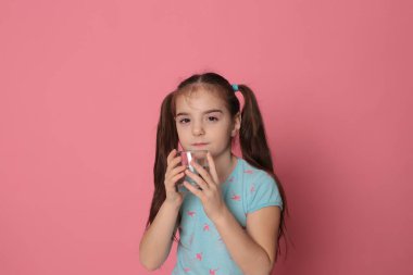 A beautiful girl of eight years drinks water from a transparent glass on a pink background in a blue T-shirt