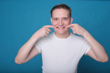 Beyaz tişörtlü komik, yakışıklı genç bir adamın şık portresi. Parlak mavi arka planda mükemmel bir ruh hali içinde.