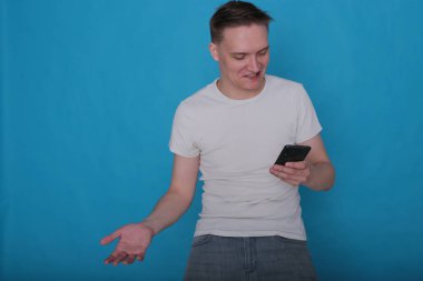 Beyaz tişörtlü komik, yakışıklı genç bir adamın şık portresi. Parlak mavi arka planda mükemmel bir ruh hali içinde.