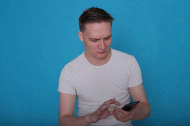 Beyaz tişörtlü komik, yakışıklı genç bir adamın şık portresi. Parlak mavi arka planda mükemmel bir ruh hali içinde.