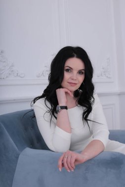 30 yıllık mutlu bir kadın, kıvırcık siyah saçlı ve beyaz elbiseli bir gül mavi koltukta oturuyor.