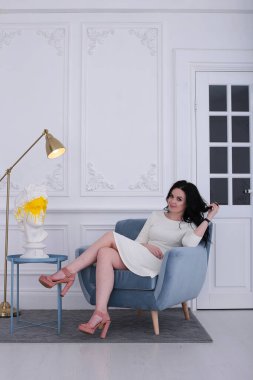 30 yıllık mutlu bir kadın, kıvırcık siyah saçlı ve beyaz elbiseli bir gül mavi koltukta oturuyor.