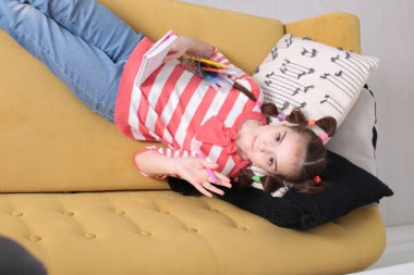 Sekiz yaşında, keyifli bir kız çizgili bir kazakla komik bir saç stili ile kahverengi bir kanepeye oturmuş elinde bir defter ve elinde keçeli kalemle.