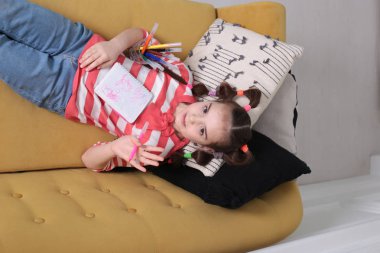 Sekiz yaşında, keyifli bir kız çizgili bir kazakla komik bir saç stili ile kahverengi bir kanepeye oturmuş elinde bir defter ve elinde keçeli kalemle.