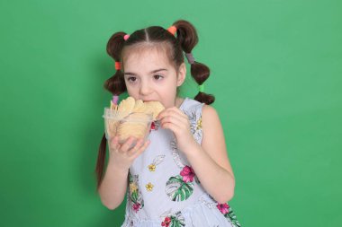 Sekiz yaşında güzel bir kız, komik saç stiliyle büyük mavi bir koltukta oturuyor ve parlak yeşil bir arka planda patates cipsi yiyor.